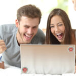 Fröhliches junges Paar, das auf einem Bett vor einem Laptop sitzt und SHIELD EMF-Schutzprodukte verwendet, um sich vor elektromagnetischer Strahlung zu schützen, glückliche Nutzer von EMF-Schutz.