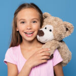 Hübsches Mädchen mit Teddybär, Kinder, Spielzeug, Lächeln, kindgerecht, kindliches Gesicht, Freude, Spaß, kindliches Glück, unschuldiges Lächeln, Flugzeug, Spielzeit, kindgerechte Kleidung, pinkes T-Shirt, freundliche Atmosphäre, Kinderliebe, kindliche Unbeschwertheit, kindliche Freude, kindliche Gesichter, positive Stimmung.