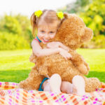 Kindermädchen umarmt großen Plüschbär im Freien bei sonnigem Wetter, symbolisiert kindliche Geborgenheit und Schutz vor elektromagnetischer Strahlung.