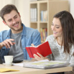 Zwei Unternehmer arbeiten an einem Tisch, einer liest in einem roten Notizbuch, während der andere an einem Laptop arbeitet, umgeben von Dokumenten, mit Fokus auf EMF-Schutz.