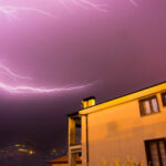 Lightning strike during a nocturnal thunderstorm over a residential house, with a dark sky and electrical discharge.